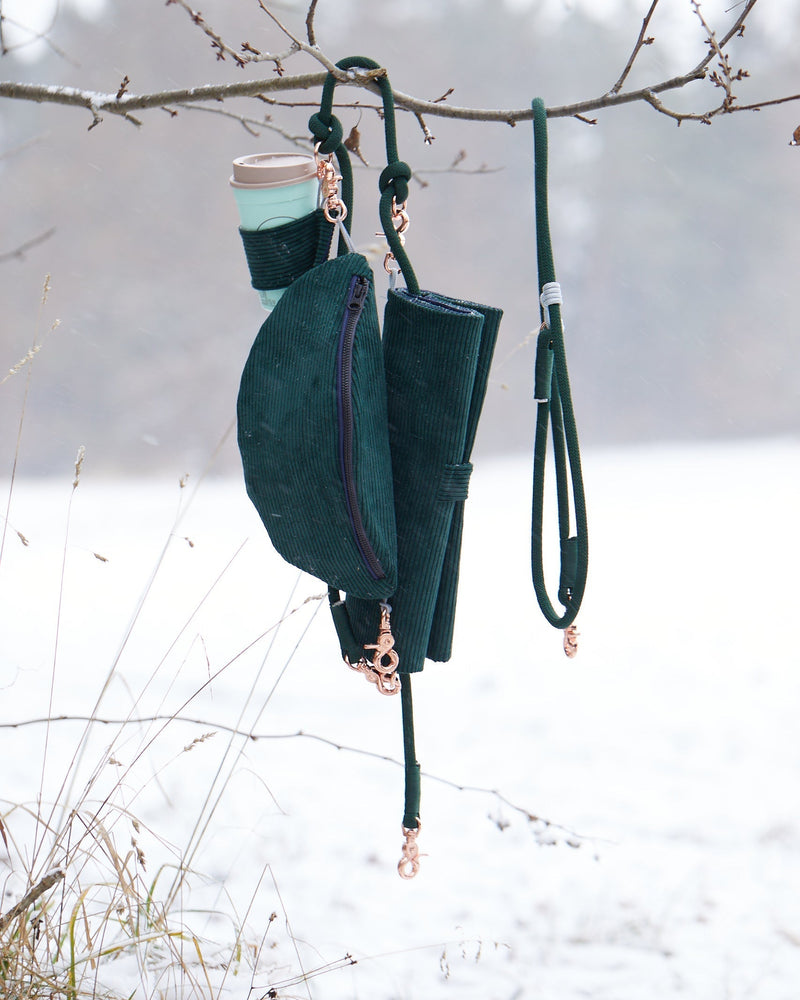 Foxc Gassi-Set bestehend aus einer grünen Cord-Bauchtasche mit Becherhalter, einer passenden Hundedecke und einer grünen Click-On-Leine, an einem Ast aufgehängt. Im Hintergrund ist eine schneebedeckte Landschaft sichtbar.