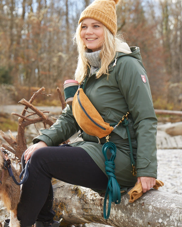 Frau mit einem Gassi-Taschen-Set in Oker, getragen als Bauchtasche. Sie sitzt auf einem Baumstamm in einem herbstlichen Wald und hält eine grüne Schleppleine sowie ein paar Accessoires für Hunde.