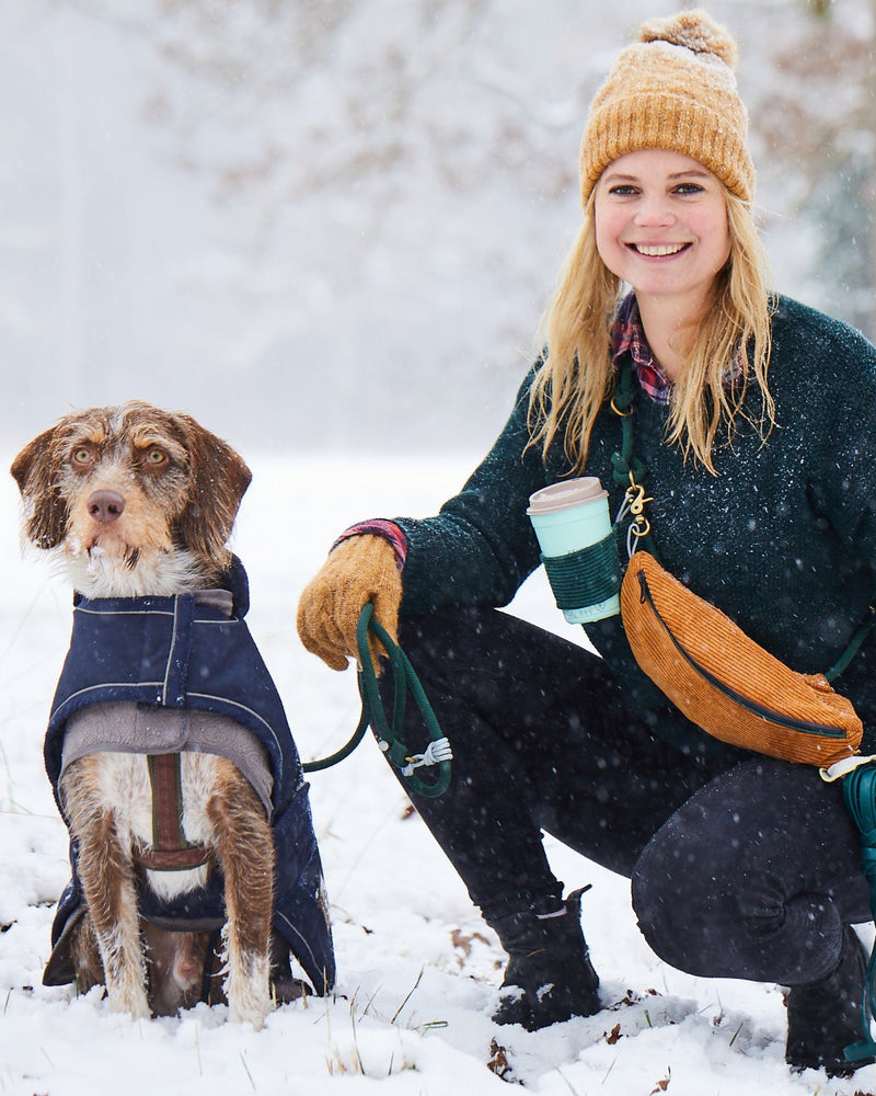 Frau mit Hund im Schnee, ausgestattet mit dem praktischen Foxc.Shop Gassitaschen-Set. Das Set umfasst einen Becherhalter mit Kaffee und eine stilvolle Tasche in ocker, goldbraun, ideal für winterliche Spaziergänge. Perfekte Kombination aus Funktionalität und Design für Hundebesitzer.